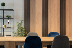 Modern conference/dining area with a long wooden table, blue and gray chairs, a vertical wooden slat wall, and a black metal shelf with plants on the left.